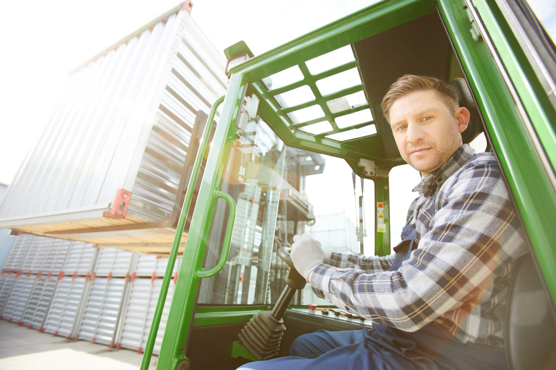 Serious handsome brutal forklift driver with stubble holding steering wheel and looking at camera while working at cargo area