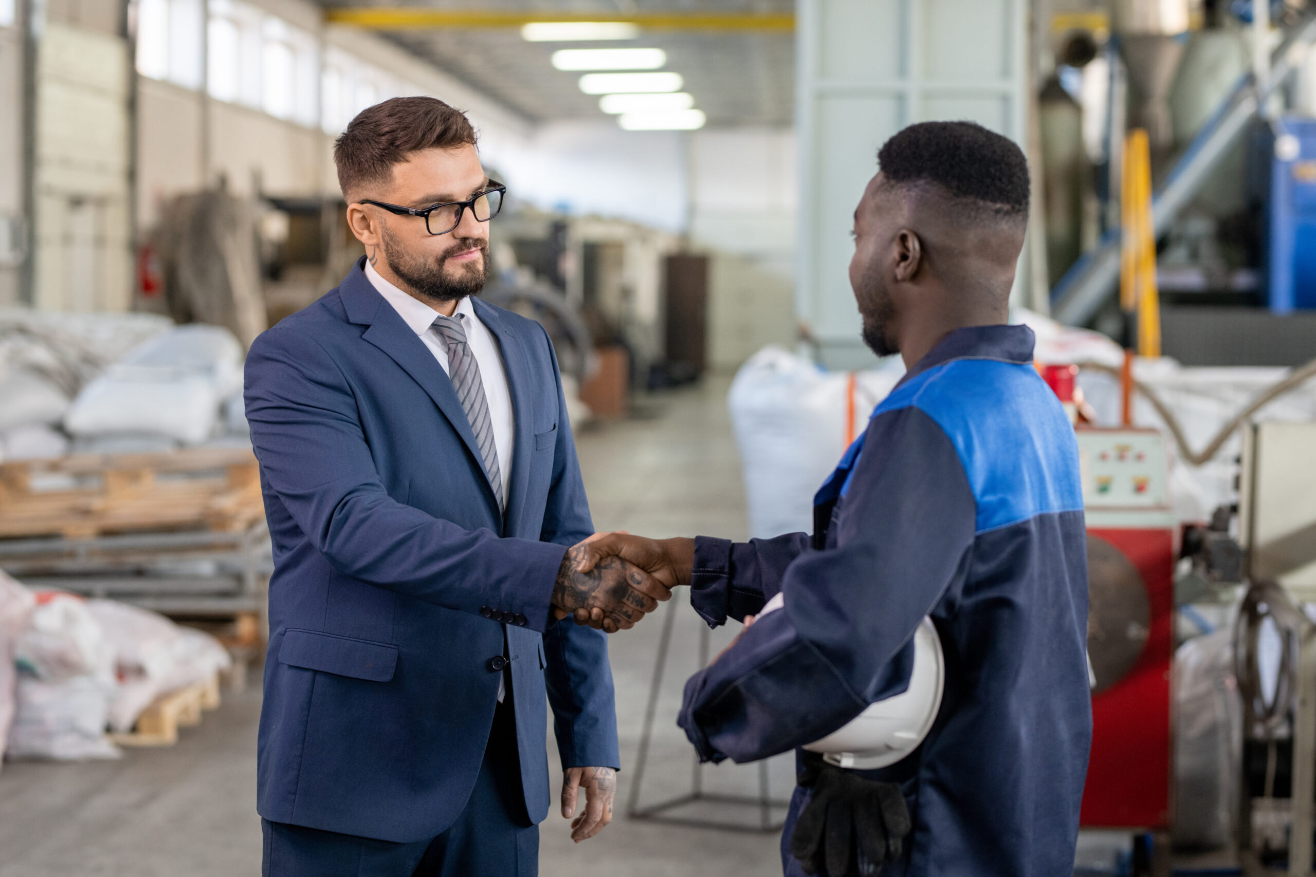 Young elegant businessman in suit and eyeglasses shaking hand of African worker of large industrial plant after making deal and signing contract