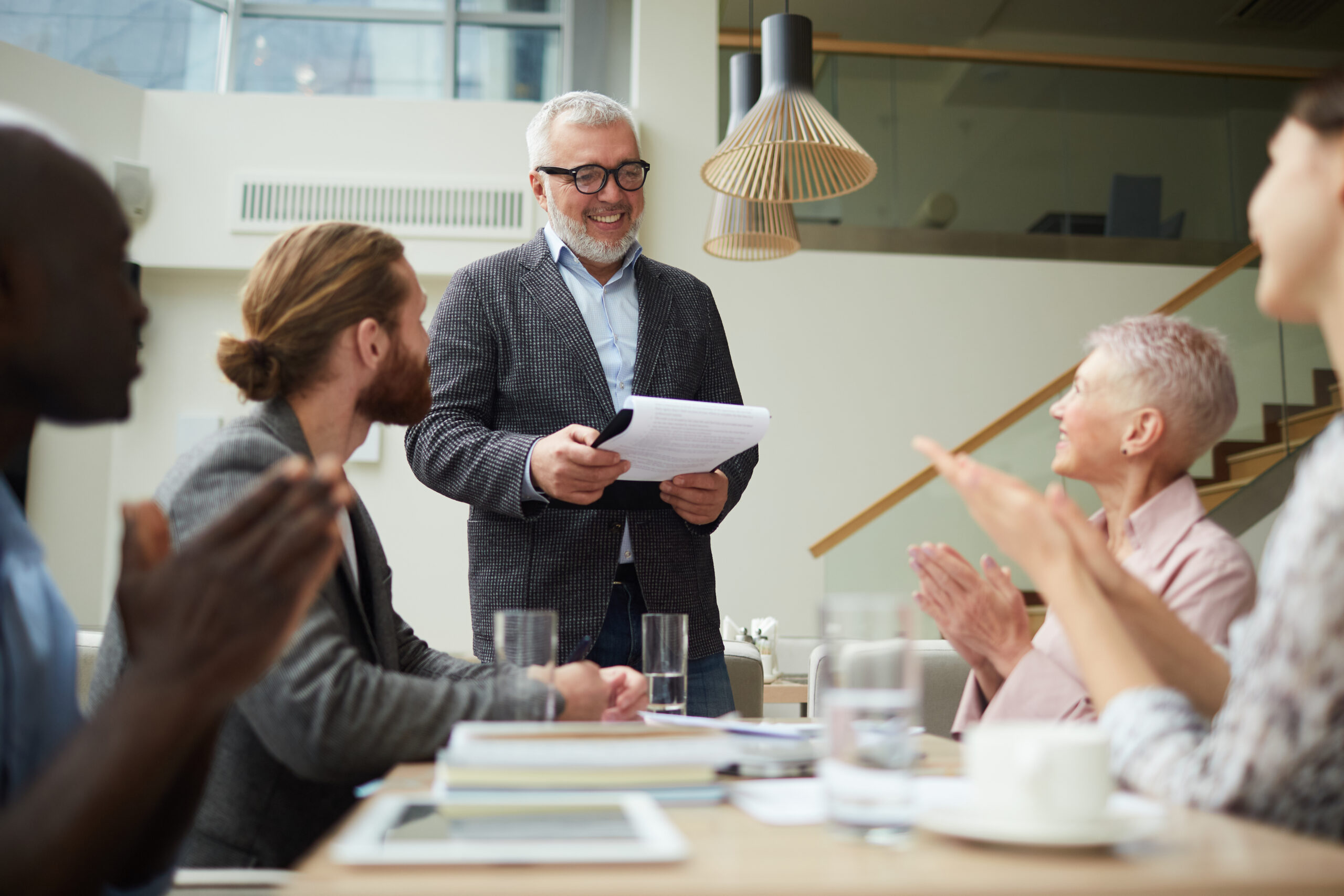 Portrait of smiling senior businessman leading meeting with employees in office and holding clipboard, copy space