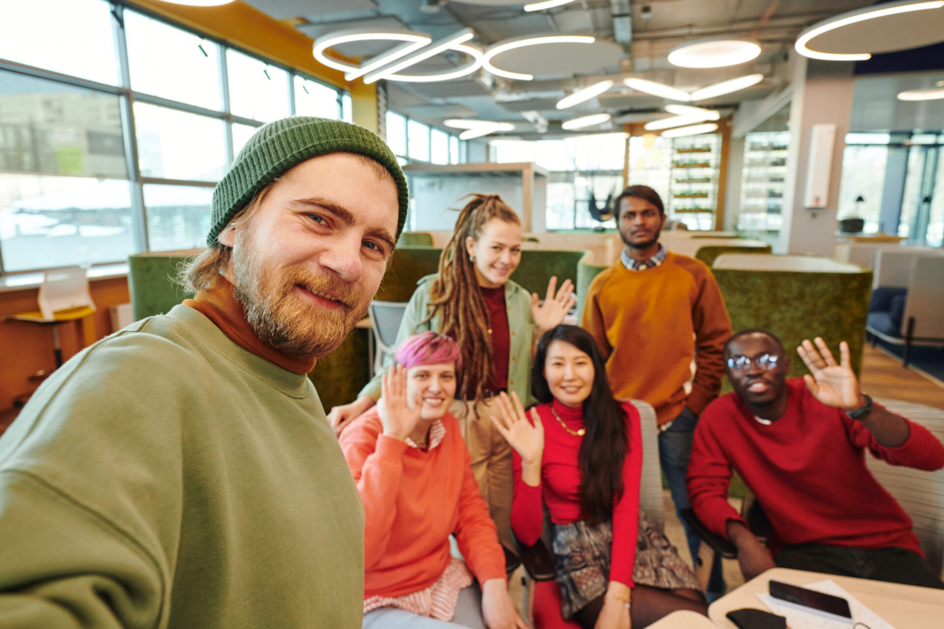 Happy young bearded businessman in casualwear making selfie with his cheerful intercultural colleagues sitting by workplace on background