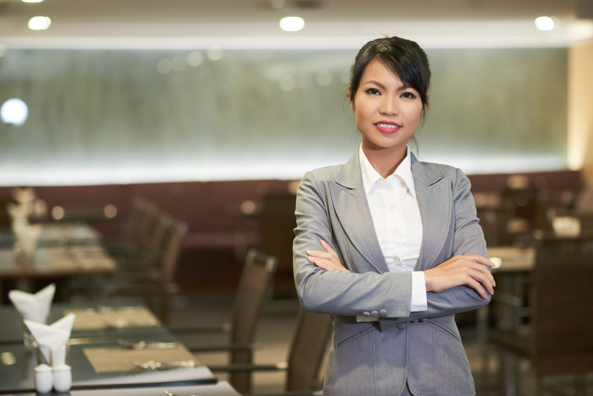 Smiling elegant Asian woman in suit working as hostess in hotel restaurant looking at camera