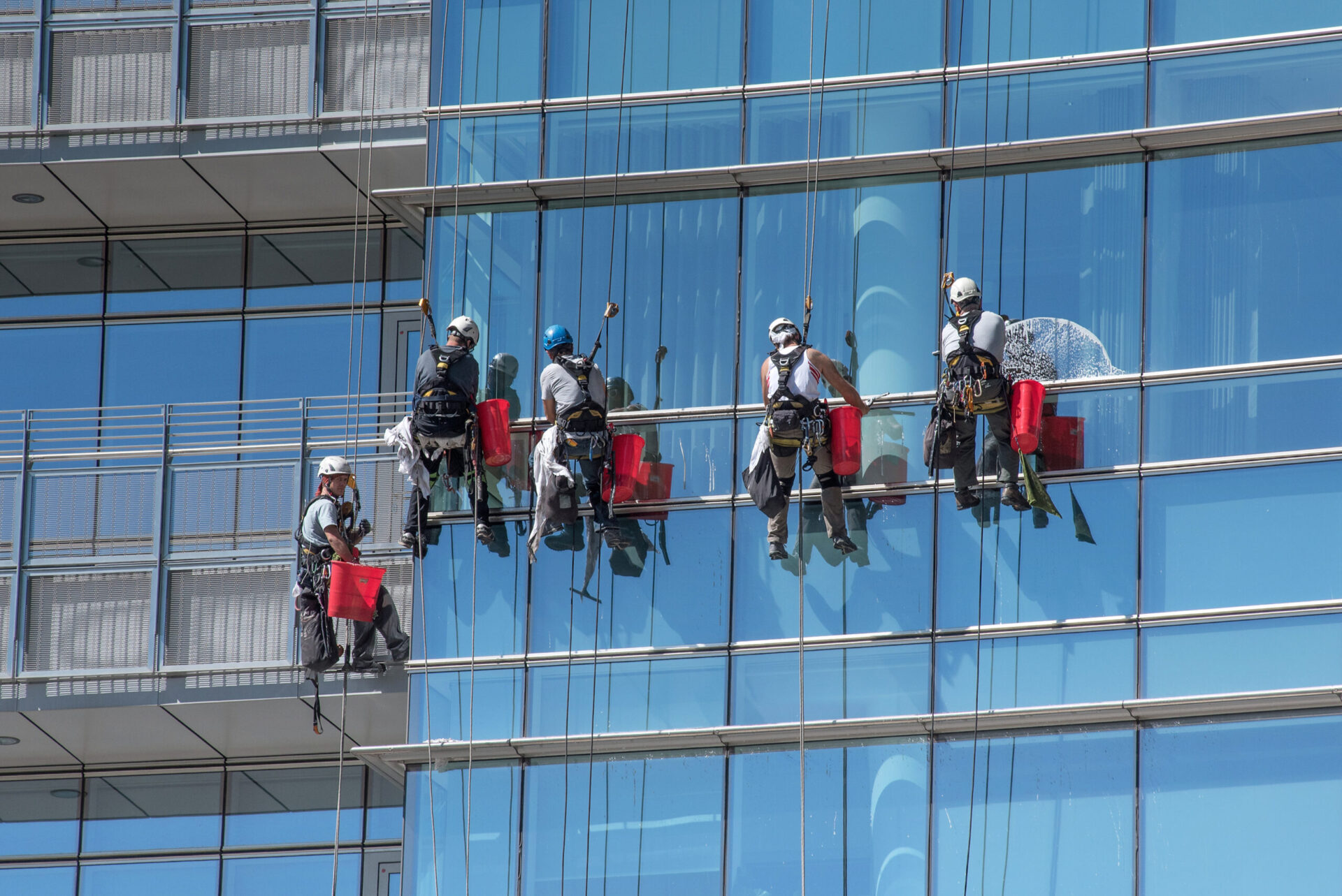 Group of window cleaners suspended from cables with safety harnesses cleaning the external glass facade of a modern skyscraper