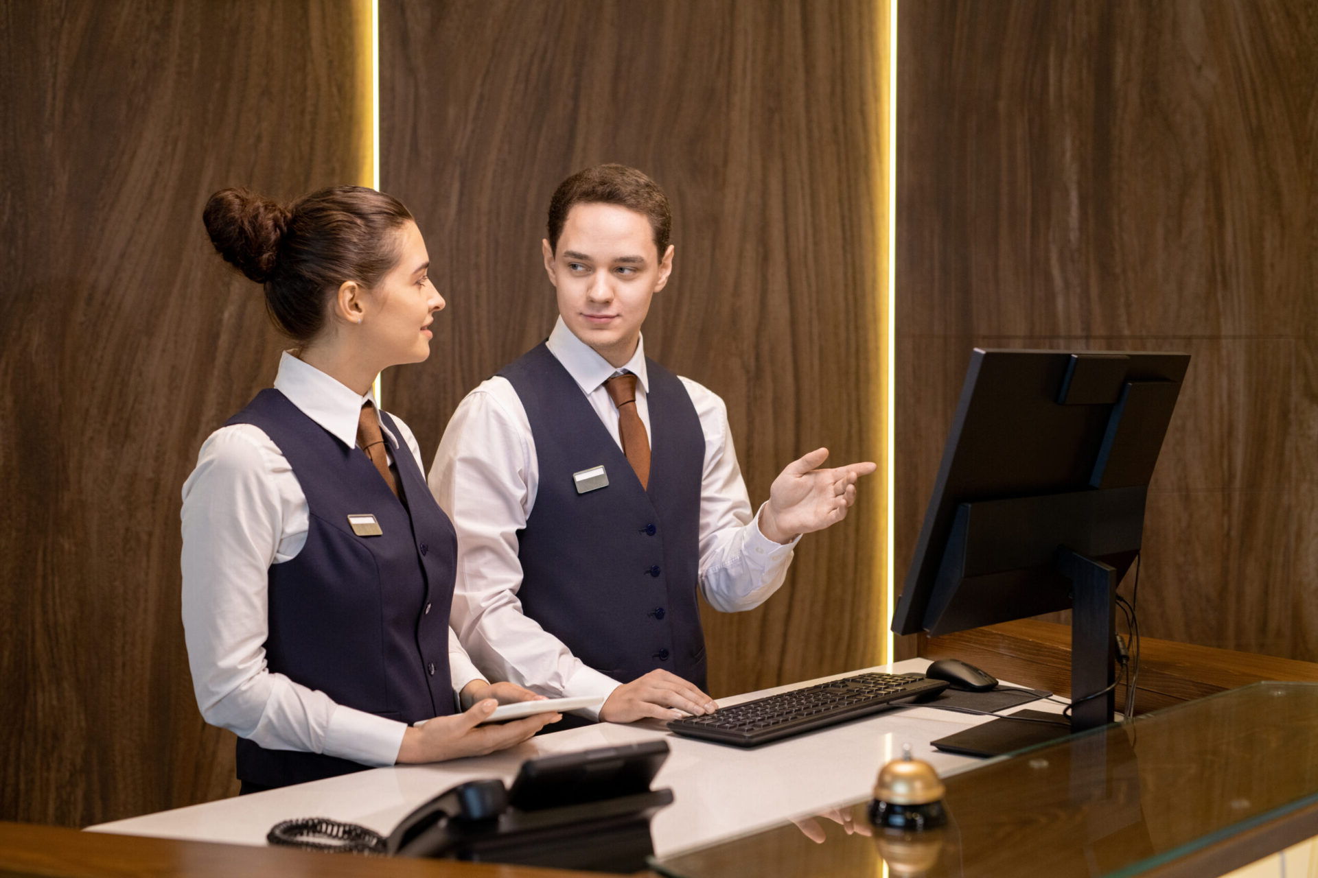 One of two young hotel receptionists standing by counter, looking at colleague with touchpad and pointing at computer screen